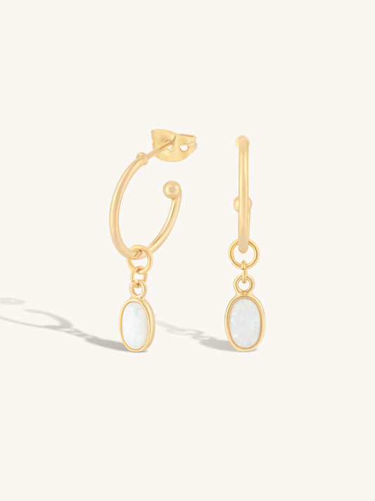 A pair of gold hoop earrings with oval white opal-like dangles, shown on a clean off-white background with soft shadows.