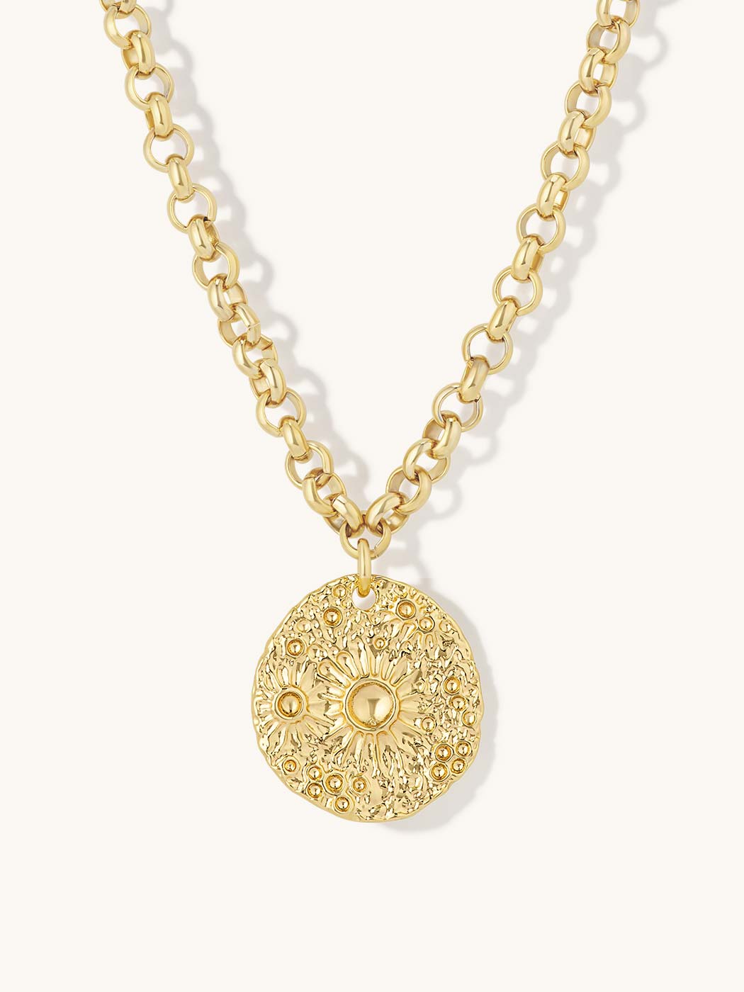 A gold chain necklace featuring a textured, circular medallion pendant with a sunburst floral pattern, shown against a light cream background.