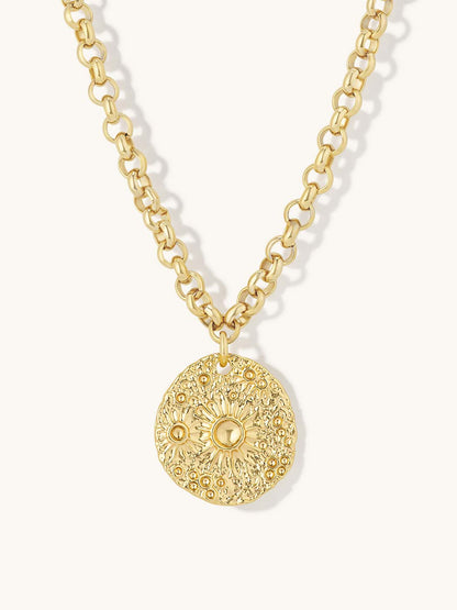 A gold chain necklace featuring a textured, circular medallion pendant with a sunburst floral pattern, shown against a light cream background.