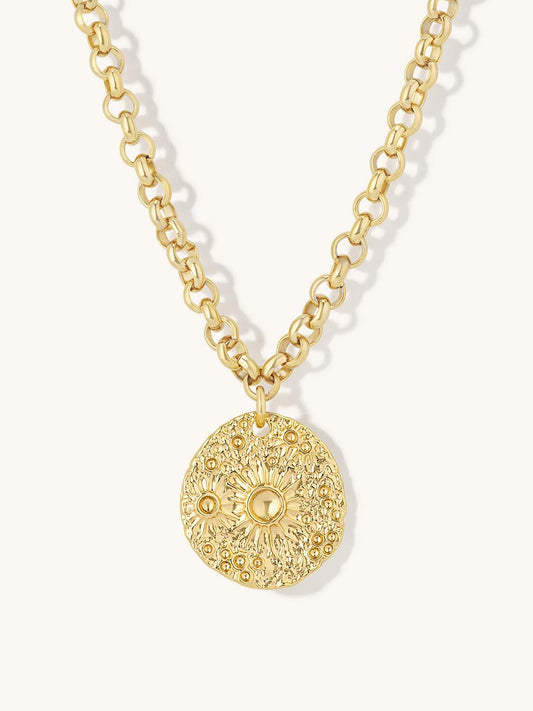 A gold chain necklace featuring a textured, circular medallion pendant with a sunburst floral pattern, shown against a light cream background.