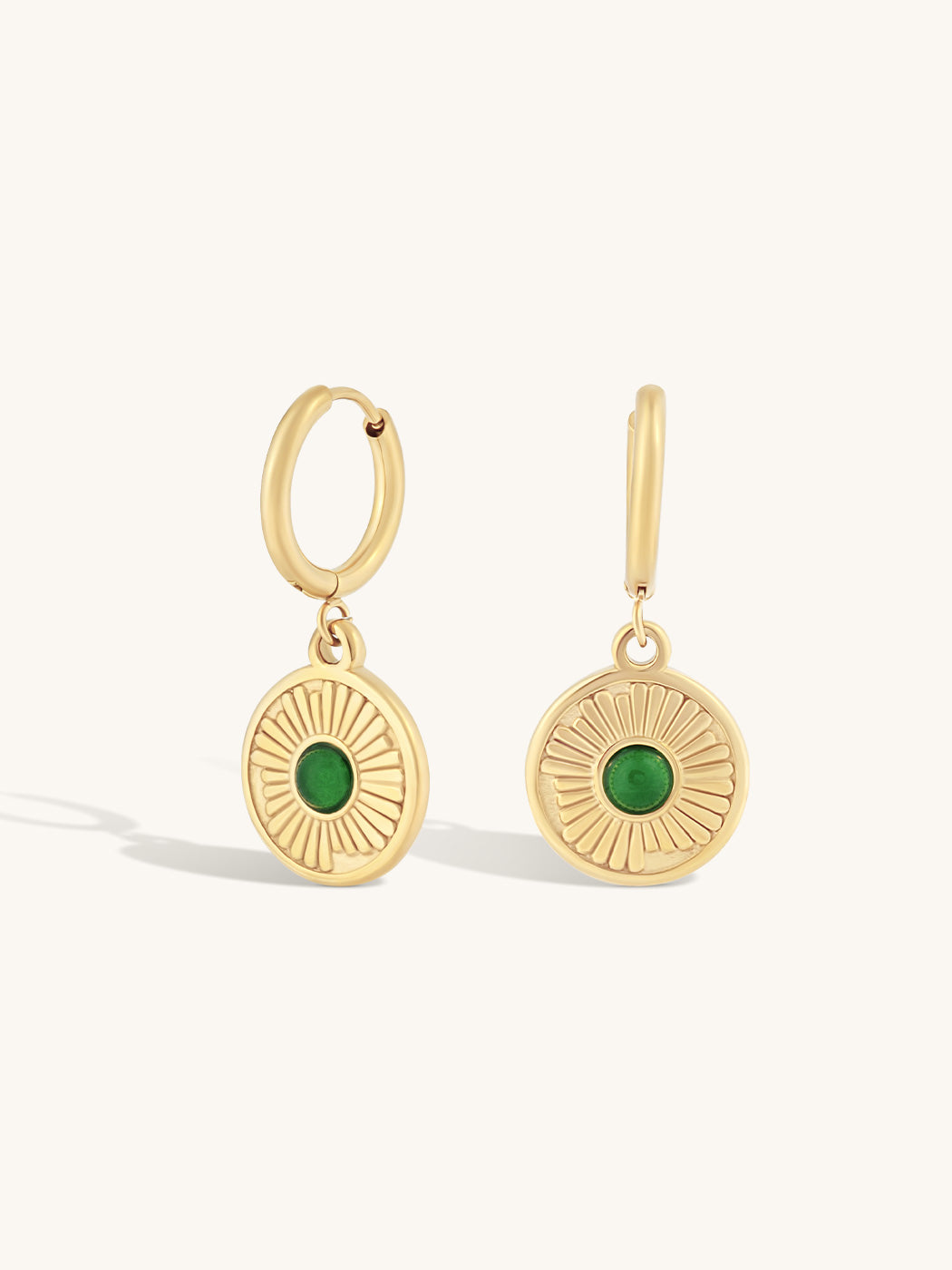 A pair of gold hoop earrings with circular sunburst disc charms featuring green oval stones, shown on a light cream background.