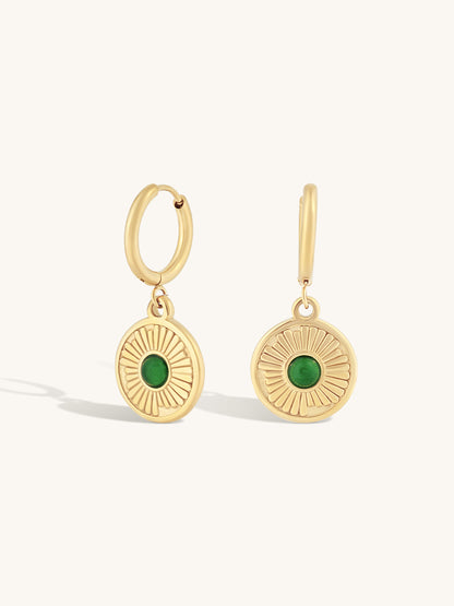 A pair of gold hoop earrings with circular sunburst disc charms featuring green oval stones, shown on a light cream background.