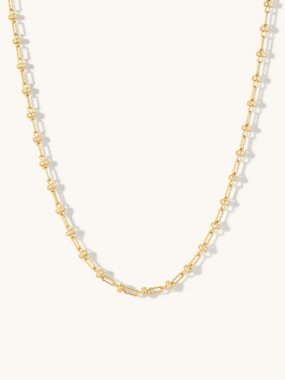 A delicate gold chain necklace featuring repeating small, textured oval links, displayed against a clean off-white background.
