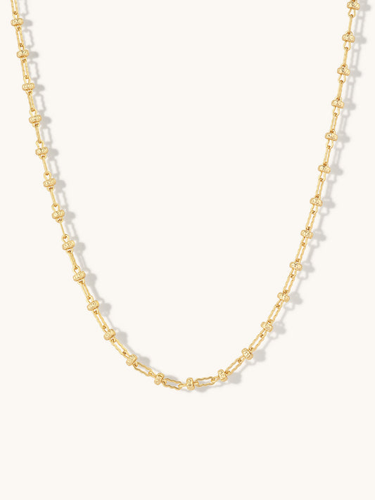 A delicate gold chain necklace featuring repeating small, textured oval links, displayed against a clean off-white background.