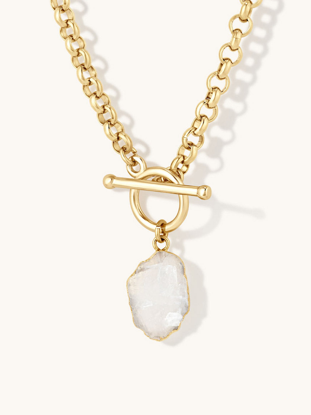 A gold chain necklace with a toggle clasp and a raw, irregular moonstone gemstone pendant, shown on a light cream background.