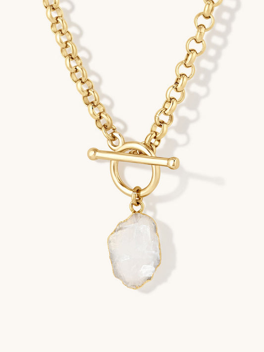 A gold chain necklace with a toggle clasp and a raw, irregular moonstone gemstone pendant, shown on a light cream background.