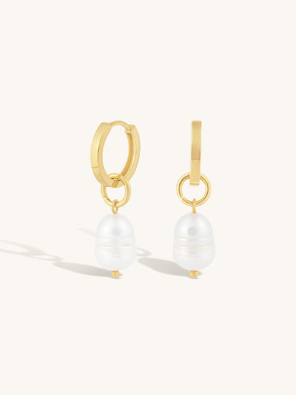 A pair of gold huggie hoop earrings with a dangling white baroque pearl, shown on a clean off-white background.