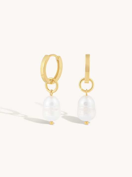 A pair of gold huggie hoop earrings with a dangling white baroque pearl, shown on a clean off-white background.