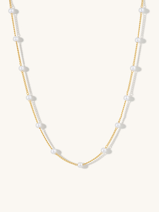 A delicate gold chain necklace featuring evenly spaced, small white pearl beads, displayed against a clean, off-white background.