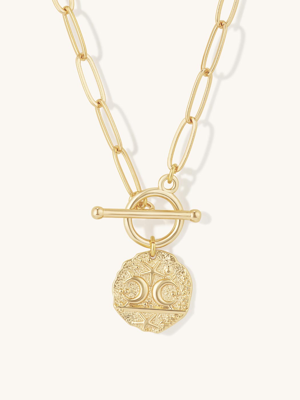 A gold-tone chain necklace featuring a toggle clasp and a circular coin-style pendant with engraved celestial details, shown against a light off-white background.