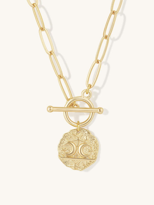 A gold-tone chain necklace featuring a toggle clasp and a circular coin-style pendant with engraved celestial details, shown against a light off-white background.