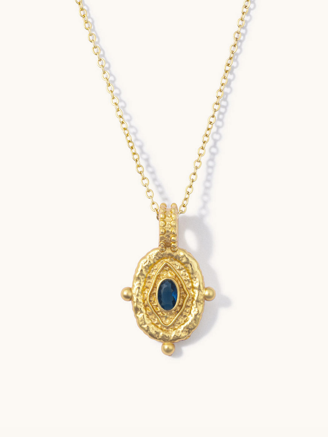 A gold pendant necklace featuring a textured oval design with a central dark blue oval stone, displayed on a clean off-white background.