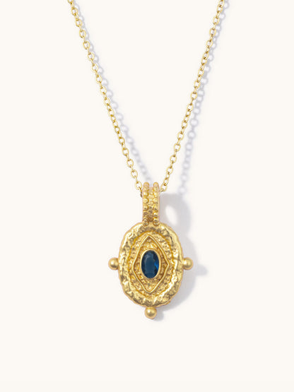 A gold pendant necklace featuring a textured oval design with a central dark blue oval stone, displayed on a clean off-white background.
