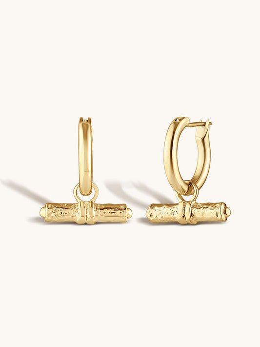 A pair of gold huggie hoop earrings with textured horizontal bar charms, displayed on a clean off-white background.