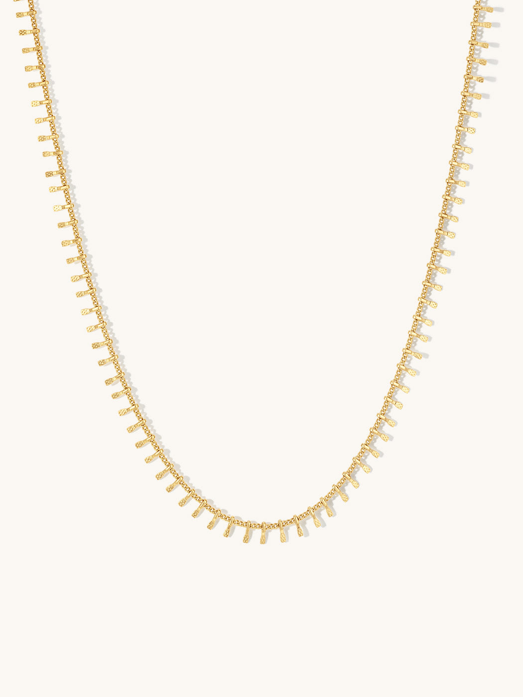 A delicate gold chain necklace featuring small, regularly spaced rectangular drop charms, displayed against a solid off-white background.