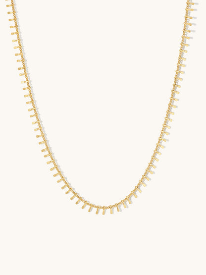 A delicate gold chain necklace featuring small, regularly spaced rectangular drop charms, displayed against a solid off-white background.