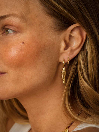 Close-up of a woman's ear wearing a small gold fish-shaped dangle earring and a tiny gold hoop earring, with blonde hair and warm natural lighting.