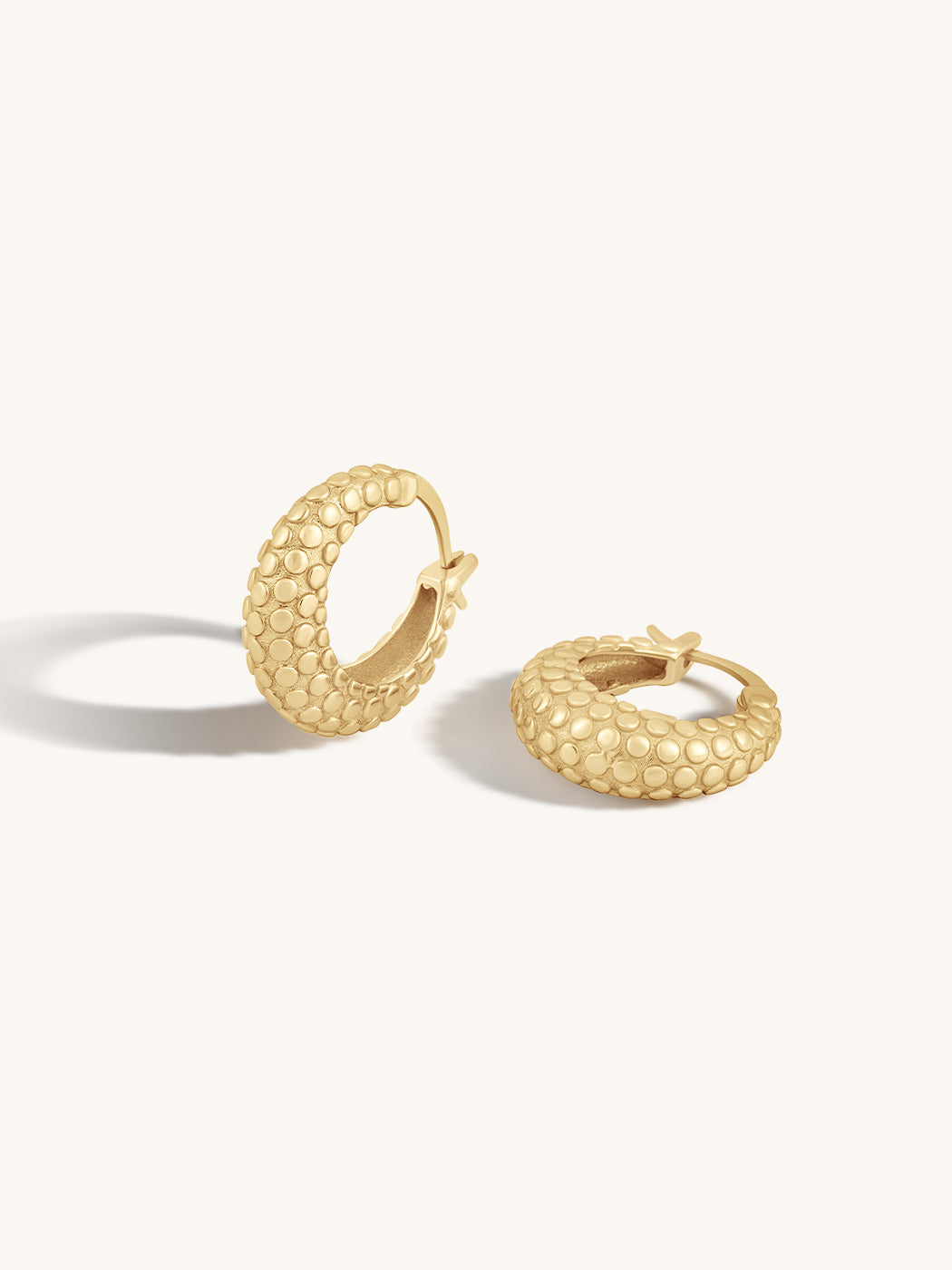 A pair of textured gold hoop earrings with a beaded, dimpled surface finish, displayed on a clean off-white background.