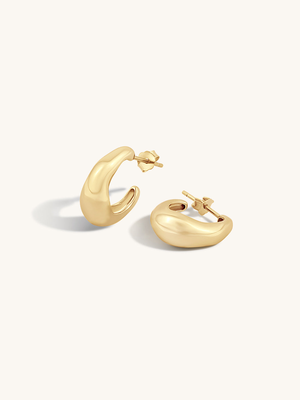 A pair of polished gold huggie hoop earrings with a rounded, organic shape, displayed on a clean off-white background.